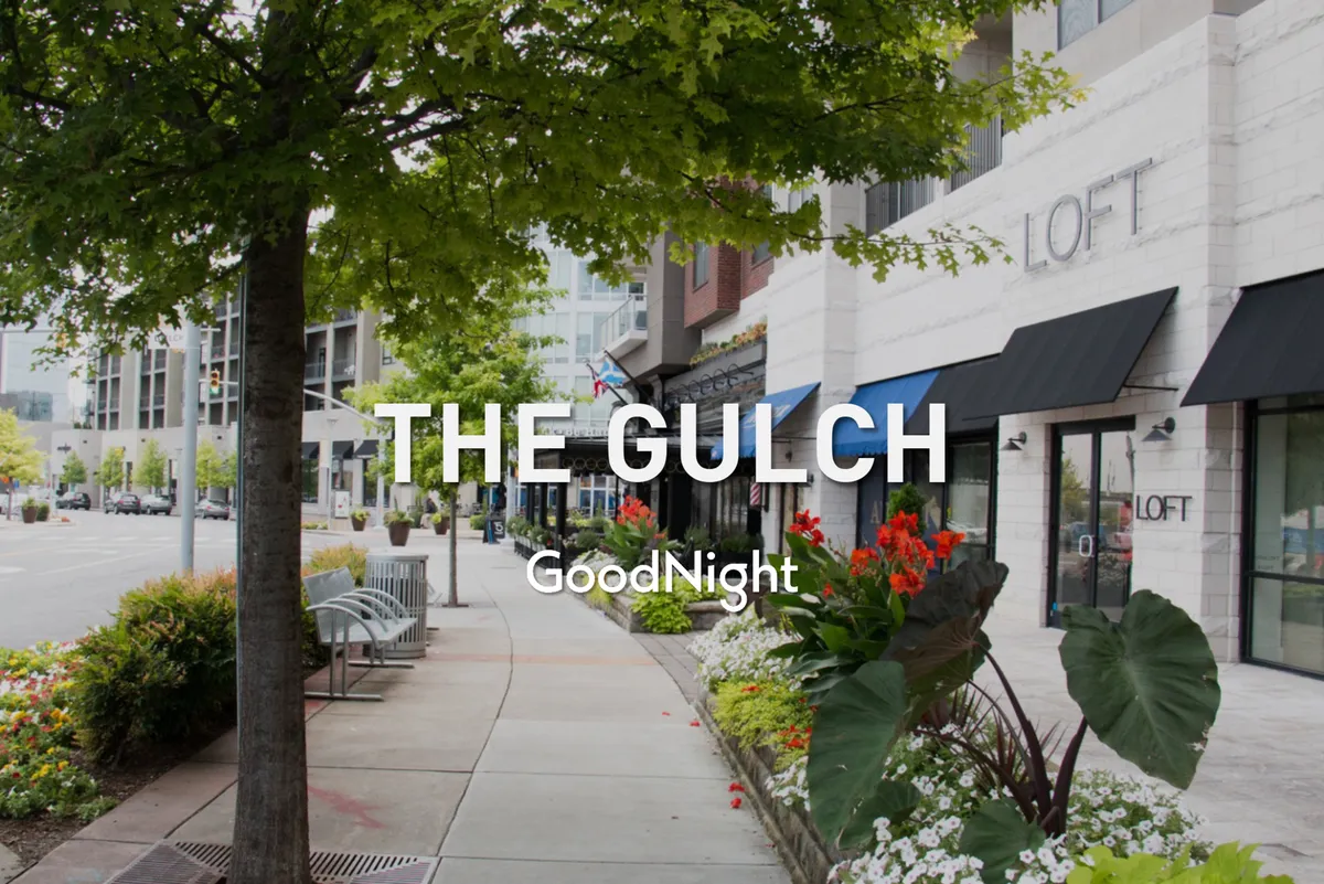 A lively exterior scene showcasing tree-lined sidewalks, benches, colorful flower beds, and storefronts, inviting residents to enjoy a walkable neighborhood atmosphere. The area looks clean and vibrant, suggesting a pet-friendly environment ideal for daily strolls with pets and socializing with neighbors.