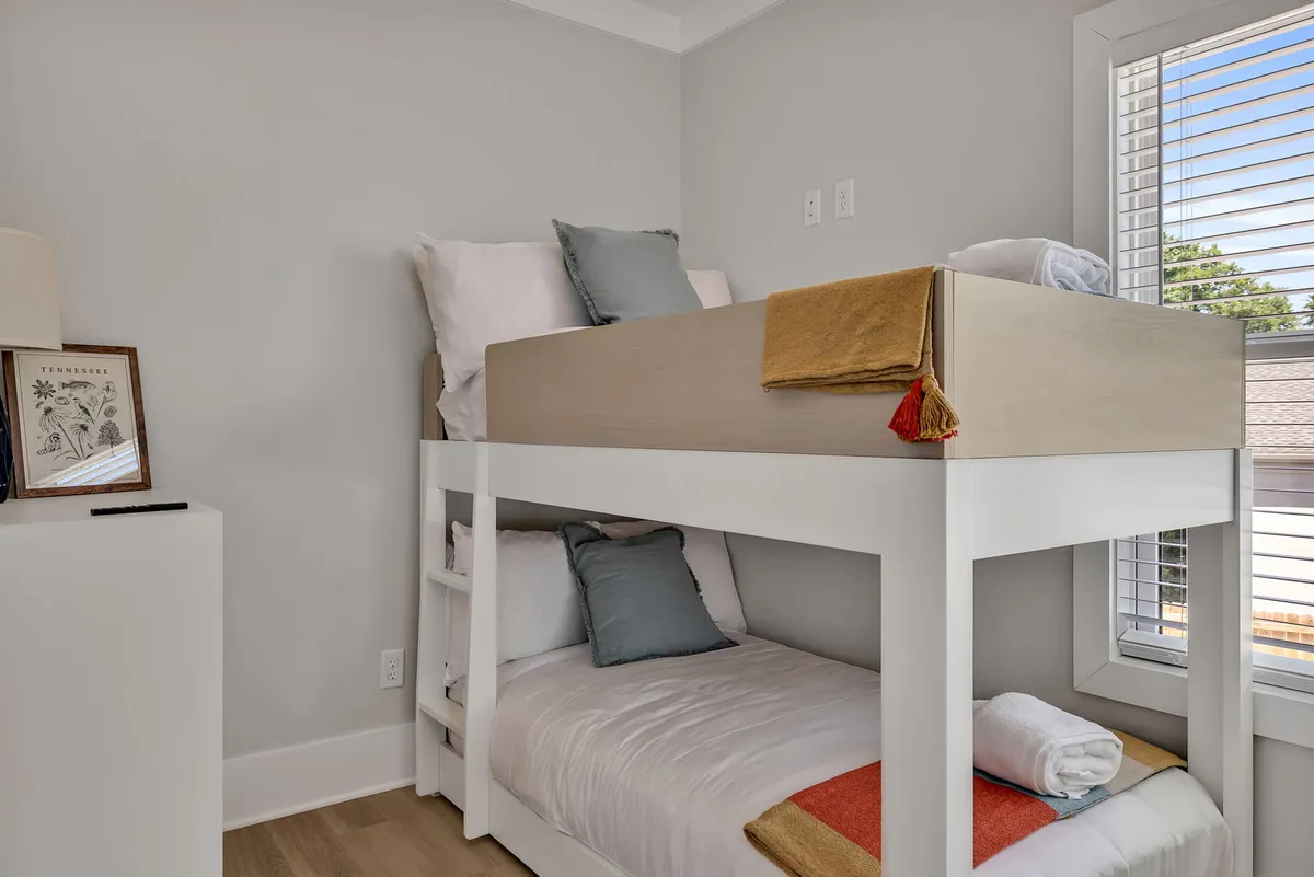 A cheerful fourth bedroom with bunk beds draped in decorative pillows, a window with blinds letting in natural light, and a playful atmosphere. The setup is welcoming for families and children.