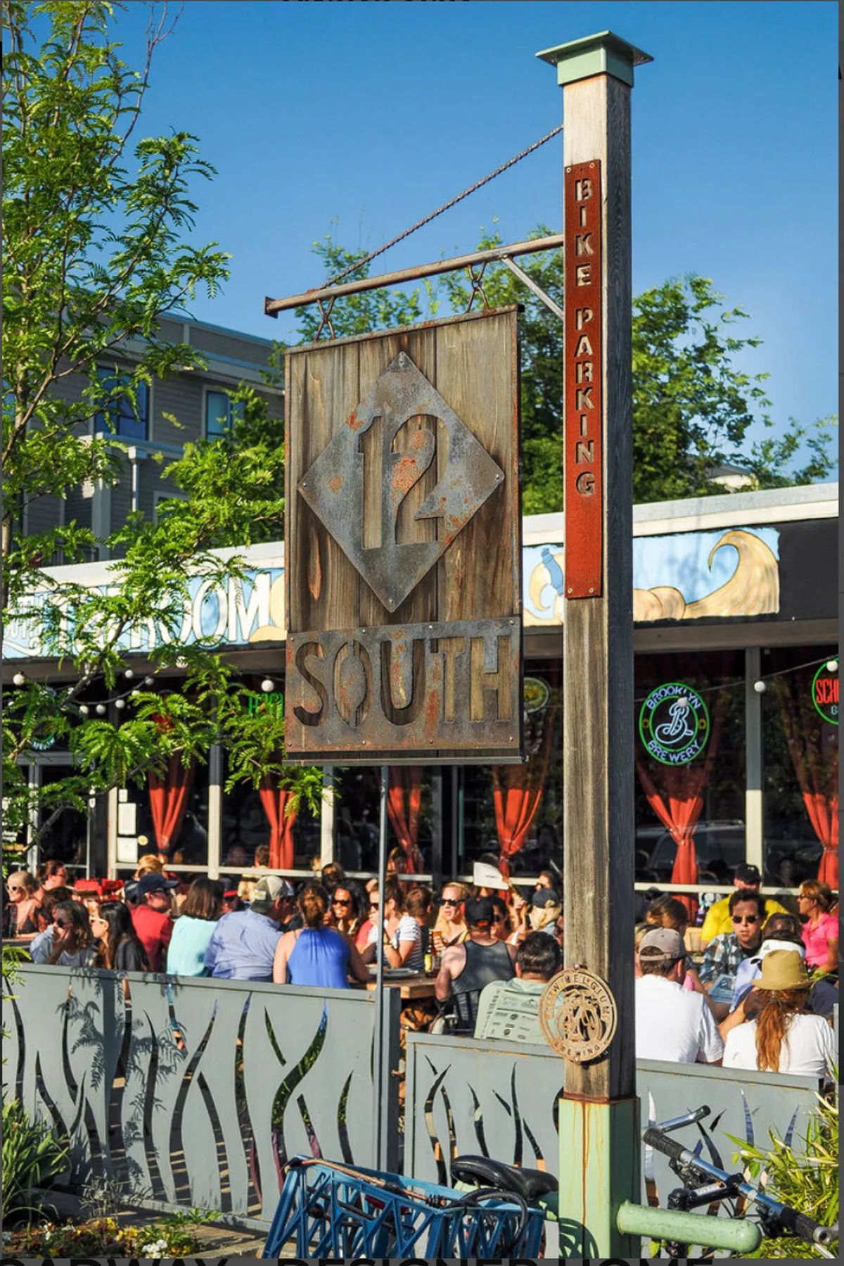 Crowds gather around outdoor tables beneath the 12 South sign, with a bustling, energetic street scene full of diners, bikers, and community members. There is a sense of communal enjoyment and vibrant local energy.