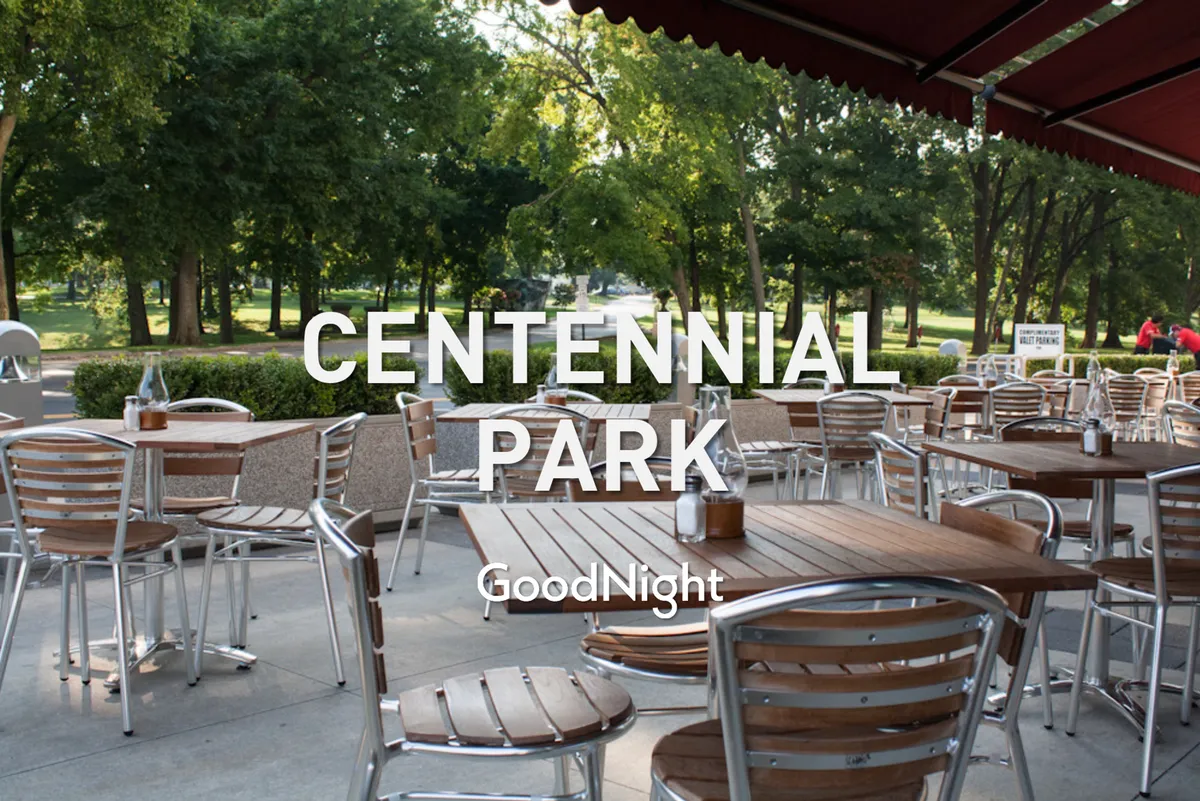 An outdoor dining area with wooden tables, green trees, and a park pathway, evoking a relaxed, family-friendly atmosphere ideal for casual meals and social gatherings in nature.