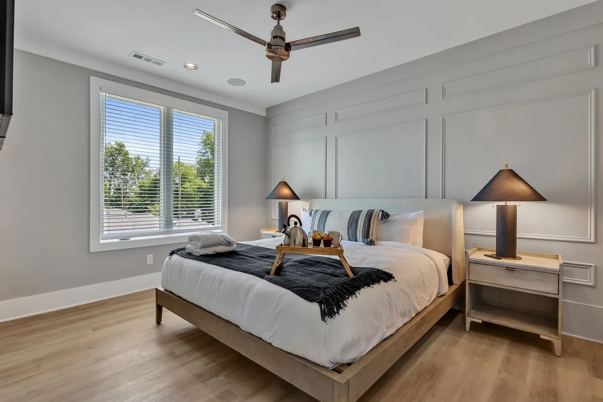 A serene bedroom featuring a queen-sized bed with a decorative throw blanket, nightstands with warm lamps on either side, and large windows with adjustable blinds allowing for plenty of natural light. The overall mood is tranquil and cozy, perfect for a restful night or a peaceful morning.