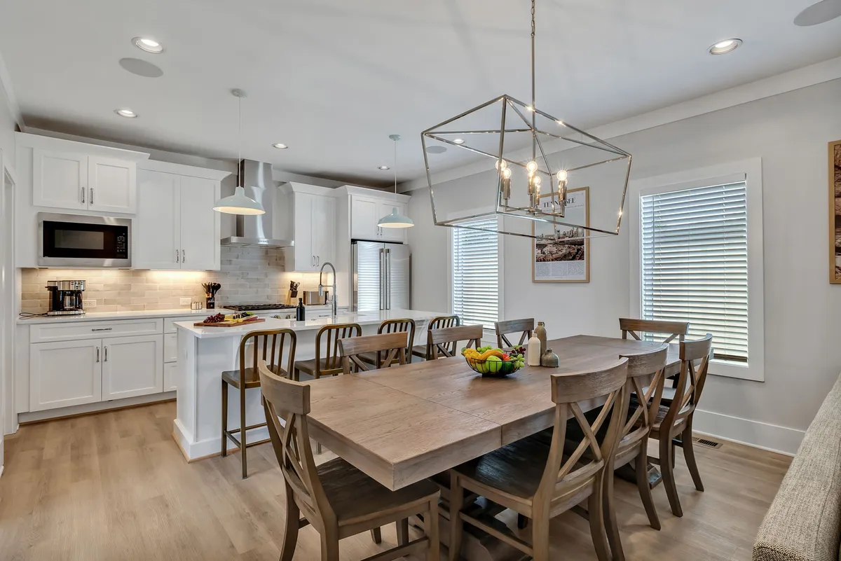 The image showcases a bright, inviting open-plan dining and kitchen area featuring a large wooden dining table under a striking geometric light fixture, complemented by a spacious kitchen island. The scene exudes warmth and offers ample space for group gatherings, meals, and celebration.
