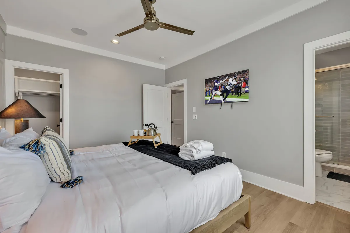 A spacious bedroom with a king bed adorned with decorative pillows, a wall-mounted Smart TV displaying a sports event, and a nightstand with a lamp and tasteful decor. The mood feels comfortable and casual, ideal for relaxing or catching up on favorite shows before bed.