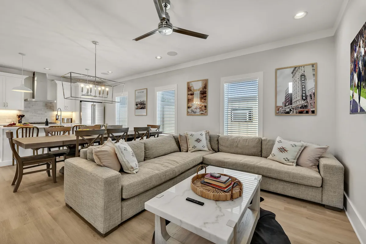 A stylish, sunlit open-concept living and dining area featuring a large sectional sofa, an elegant dining table set for eight beneath a chandelier, and curated artwork on the walls. The room exudes a sense of coziness and togetherness, ideal for group gatherings and social activities.